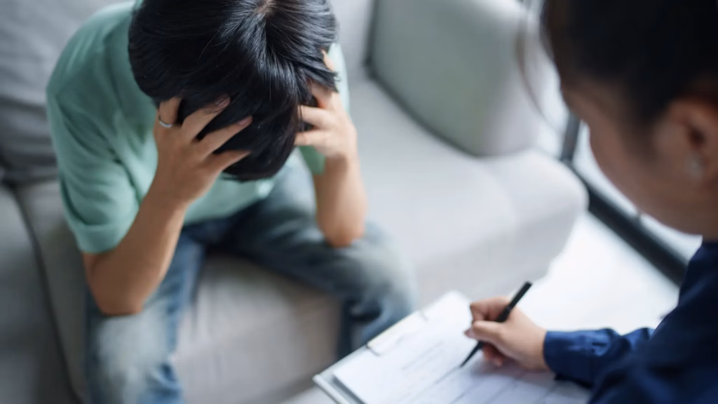 Young person sitting on a couch holding their head in distress while speaking with a therapist taking notes