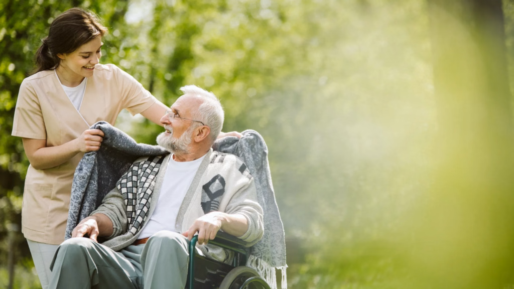 A caregiver draping a blanket over a senior man in a wheelchair outdoors.