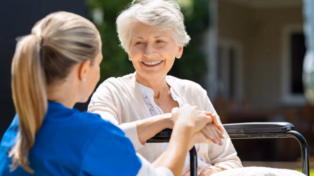 A nurse in blue scrubs holding hands and conversing with an elderly woman
